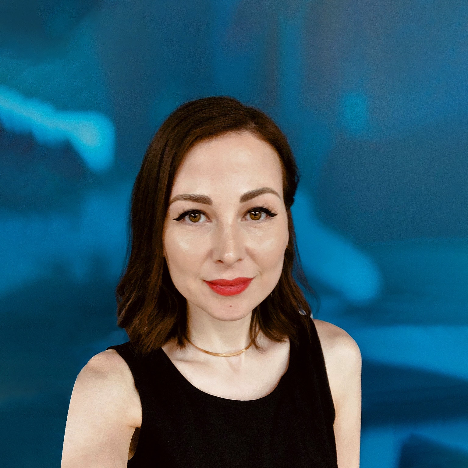 Portrait of Zeinab Hijazi with short dark-brown hair and red lipstick. She is wearing a black tank top and a gold necklace. The background is a blue and white abstract pattern.